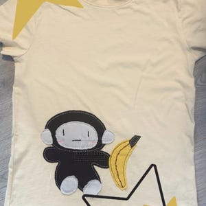 May include: A cream-coloured short-sleeve t-shirt featuring a black and white appliqué of a cartoon monkey holding a yellow banana. The shirt has a crew neckline and a size XS label printed on the inside collar. The design includes decorative stars.