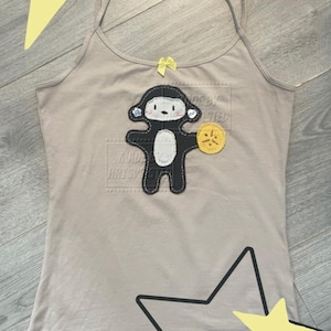 May include: A beige spaghetti strap vest top featuring a central appliqué of a black and white monkey character holding a yellow circular object. The monkey has button eyes and a small yellow bow is attached to the neckline of the vest top.