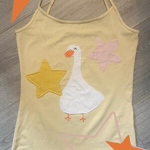 Op de afbeelding: Een lichtgeel hemdje met spaghettibandjes en een witte ganzenapplicatie met een oranje gespikkelde snavel. Het ontwerp bevat een gele ster aan de linkerkant en een lichtroze ster aan de rechterkant, met de tekst "MADE BY ARTWASTED" zichtbaar.