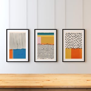 May include: Three framed abstract art prints with black and white geometric patterns and colourful accents. The prints are hung on a white wall above a wooden table.