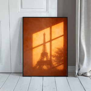 May include: Framed print featuring the Eiffel Tower in silhouette against an orange background. The image is framed in black and appears to be lit by sunlight streaming through a window, creating shadows. The print is displayed against a white door and floor.