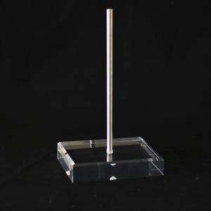 May include: A clear acrylic display stand with a rectangular base and a vertical silver metal rod. The stand is designed to showcase items and is set against a black background.