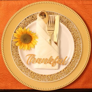 May include: A gold charger plate with a white plate on top. The white plate has a white napkin folded in the center with gold silverware and a sunflower on the side. The napkin has a wooden sign that says "Thankful" on it.