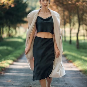 Woman wearing black silk cropped draped cowl neck camisole  and skirt outdoors with cream coat