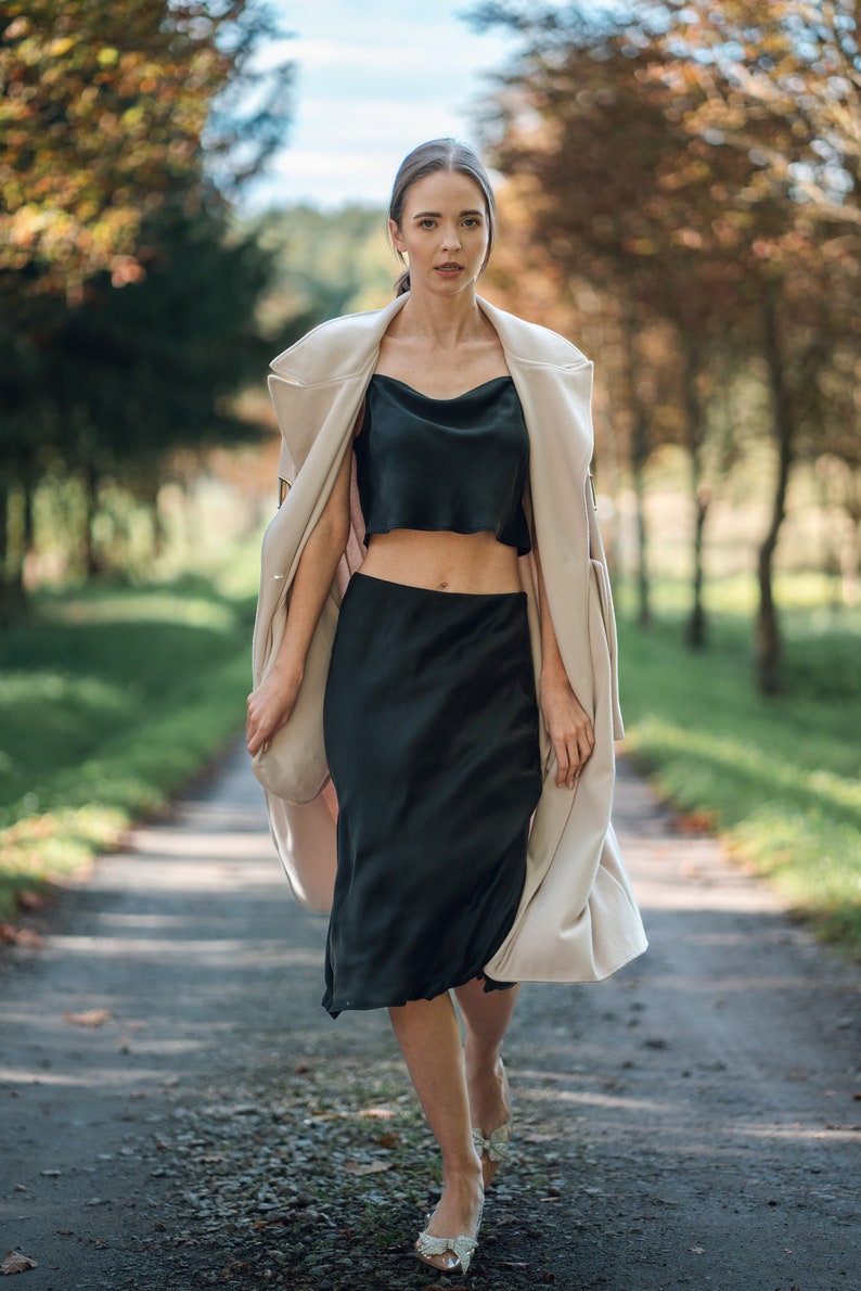 Woman wearing black silk cropped draped cowl neck camisole  and skirt outdoors with cream coat