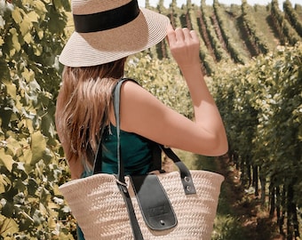 May include: A woman wearing a straw hat with a black band and a green sleeveless dress is carrying a woven straw tote bag with black leather straps and a black leather flap. The bag has a silver metal buckle.