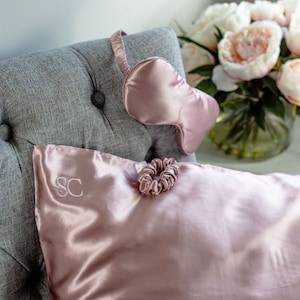 May include: A blush pink satin pillowcase with the initials "SC" embroidered on the front. A blush pink satin sleep mask and a blush pink hair scrunchie are also pictured.