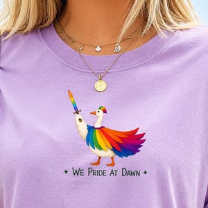 May include: A light purple long-sleeve shirt featuring a graphic of a white duck with rainbow-colored wings and a rainbow crest, holding a rainbow sword. Below the duck, the text reads: WE PRIDE AT DAWN, flanked by two small four-pointed stars.