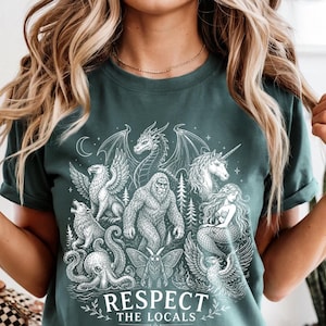 May include: A dark green t-shirt featuring a white graphic print of various mythical creatures including a dragon, unicorn, mermaid, and bigfoot. The text "RESPECT THE LOCALS" is printed in white at the bottom of the design, surrounded by stars.