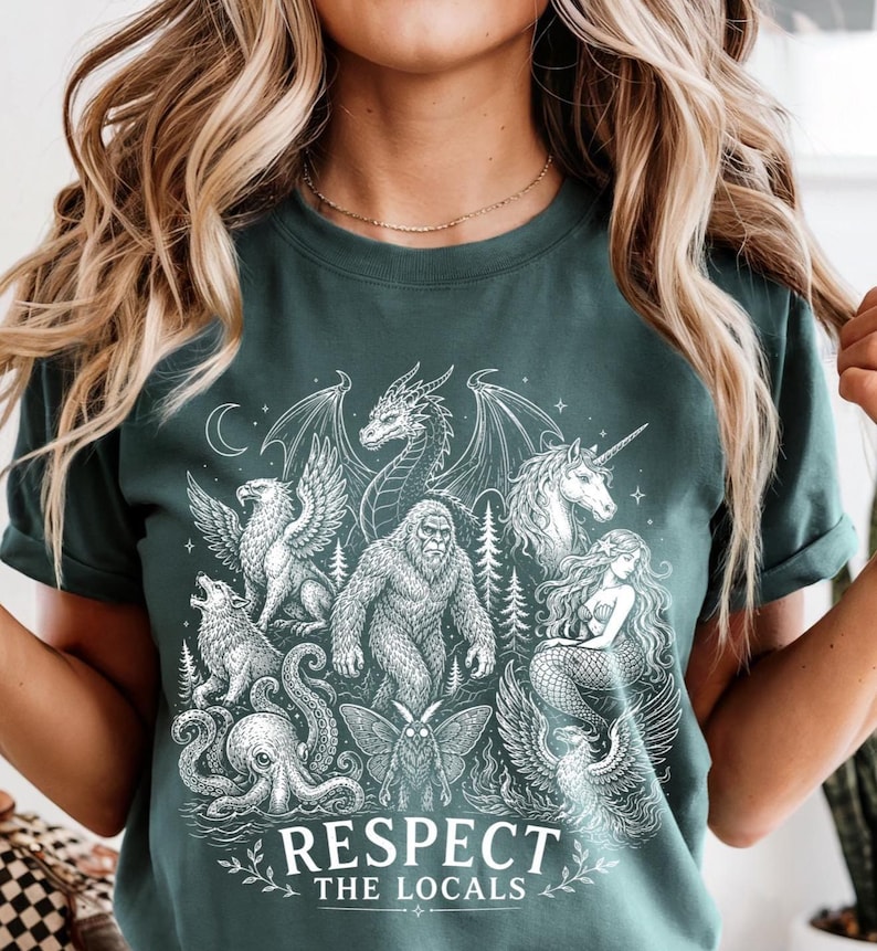 May include: A dark green t-shirt featuring a white graphic print of various mythical creatures including a dragon, unicorn, mermaid, and bigfoot. The text "RESPECT THE LOCALS" is printed in white at the bottom of the design, surrounded by stars.
