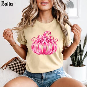 Op de afbeelding: Een lichtbeige t-shirt met een afbeelding van een pompoen gemaakt van roze linten. Het ontwerp bevat een centraal roze bewustzijnslintje bovenop de pompoen. Het woord Butter staat in zwarte letters in de linkerbovenhoek gedrukt.