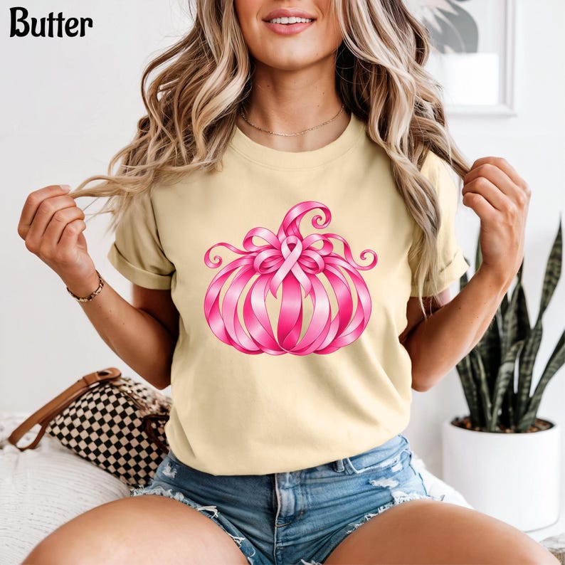 Op de afbeelding: Een lichtbeige t-shirt met een afbeelding van een pompoen gemaakt van roze linten. Het ontwerp bevat een centraal roze bewustzijnslintje bovenop de pompoen. Het woord Butter staat in zwarte letters in de linkerbovenhoek gedrukt.