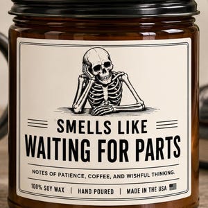 May include: A brown glass candle jar with a black metal lid and a white label. The label features a black and white illustration of a skeleton resting its head on its hand, with the text "SMELLS LIKE WAITING FOR PARTS" and "NOTES OF PATIENCE, COFFEE, AND WISHFUL THINKING. 100% SOY WAX | HAND POURED | MADE IN THE USA".