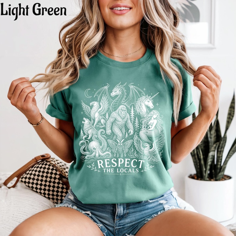 May include: A light green t-shirt featuring a white graphic print of various mythical creatures including a dragon, unicorn, mermaid, and bigfoot, with the text "RESPECT THE LOCALS" printed in a bold, white, capitalized font below the illustrations.