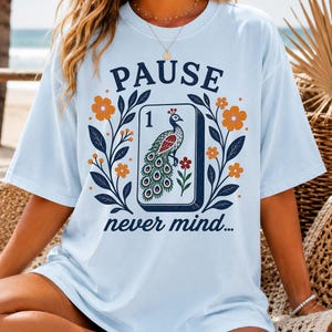 Op de afbeelding: Een lichtblauw t-shirt met een grafische print van een mahjongtegel met een pauwillustratie, het cijfer 1 en de tekst PAUSE never mind. Het ontwerp is omgeven door donkerblauwe bladeren en oranje bloemen op de voorzijde van het t-shirt.