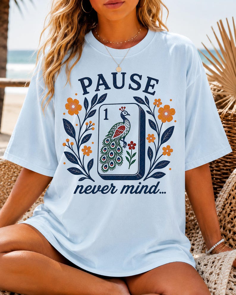 Op de afbeelding: Een lichtblauw t-shirt met een grafische print van een mahjongtegel met een pauwillustratie, het cijfer 1 en de tekst PAUSE never mind. Het ontwerp is omgeven door donkerblauwe bladeren en oranje bloemen op de voorzijde van het t-shirt.