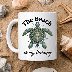 May include: White ceramic mug with a sea turtle design. The turtle is teal, green, and gold, with a mandala-style pattern. The text "The Beach is my therapy" is printed on the mug. The mug is surrounded by seashells and sand.