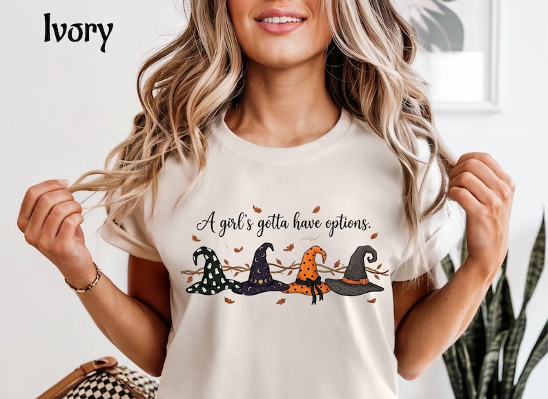 May include: An ivory t-shirt featuring a graphic print of four distinct witch hats in green, purple, orange, and black, arranged in a row with brown autumn leaves and branches. The text above the hats reads, A girl's gotta have options.