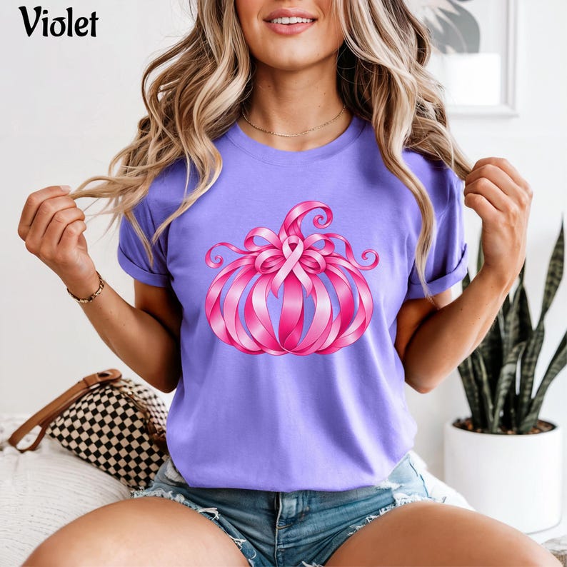Op de afbeelding: Een lichtpaars t-shirt met korte mouwen en een afbeelding van een pompoen gemaakt van roze linten. Het ontwerp bevat een centraal roze bewustzijnslintje boven op de pompoen. Het woord Violet staat in het zwart in de linkerbovenhoek gedrukt.