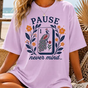 Op de afbeelding: Een lichtpaars t-shirt met korte mouwen en een grafische print van een mahjongtegel met een pauwillustratie en het cijfer 1. Het ontwerp bevat de tekst PAUSE boven de tegel en never mind... eronder, omringd door oranje bloemen en bladeren.