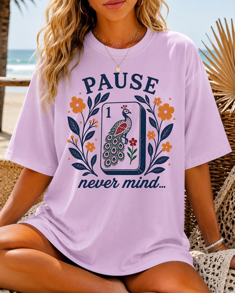 Op de afbeelding: Een lichtpaars t-shirt met korte mouwen en een grafische print van een mahjongtegel met een pauwillustratie en het cijfer 1. Het ontwerp bevat de tekst PAUSE boven de tegel en never mind... eronder, omringd door oranje bloemen en bladeren.