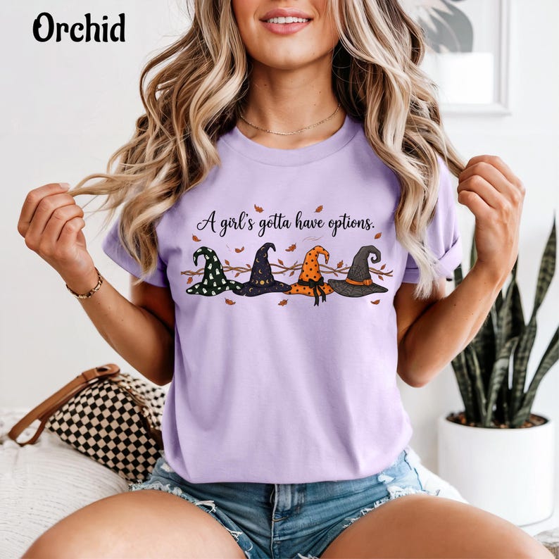 May include: A light purple t-shirt featuring the text "A girl's gotta have options." and a graphic of four distinct witch hats in green, black, orange, and dark gray, accented with small brown autumn leaves and thin branch illustrations.
