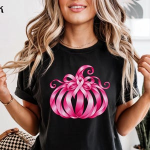 Op de afbeelding: Een zwart t-shirt met een afbeelding van een pompoen opgebouwd uit roze linten. Het woord Black staat in de linkerbovenhoek gedrukt. Het pompoenontwerp bevat een centrale roze bewustwordingslus bij het bovenste steeltje.
