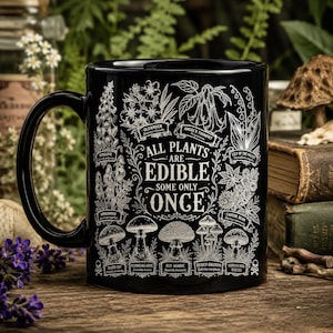 May include: A black ceramic mug featuring white botanical illustrations of various plants and mushrooms. The text reads: All plants are edible, some only once. Labeled illustrations include Oleander, Angel's Trumpet, Lily of the Valley, and Death Cap.