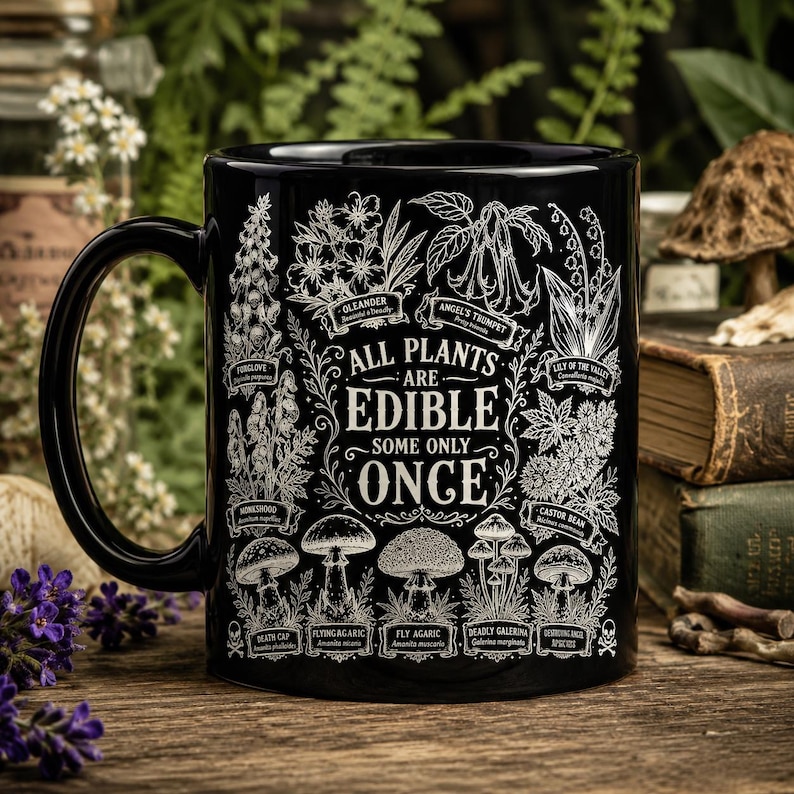 May include: A black ceramic mug featuring white botanical illustrations of various plants and mushrooms. The text reads: All plants are edible, some only once. Labeled illustrations include Oleander, Angel's Trumpet, Lily of the Valley, and Death Cap.