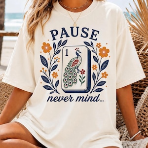 May include: An ivory t-shirt featuring a graphic print with the text PAUSE and never mind. The design includes a central rectangular tile with the number 1 and a peacock illustration, surrounded by orange flowers and dark blue leafy branches.