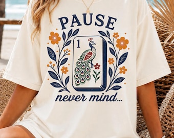 May include: An ivory t-shirt featuring a graphic print with the text PAUSE and never mind. The design includes a central rectangular tile with the number 1 and a peacock illustration, surrounded by orange flowers and dark blue leafy branches.