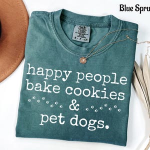 以下が含まれることがあります： 「happy people bake cookies & pet dogs.」という白い文字がプリントされた、ブルー・スプルース色の半袖Tシャツ。クルーネックで、襟の内側にはComfort Colorsのブランドラベルがあり、小さな足跡のグラフィックがデザインされています。
