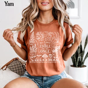 May include: A rust-colored short-sleeve t-shirt featuring a white graphic print of various plants and mushrooms. The central text reads, "ALL PLANTS ARE EDIBLE SOME ONLY ONCE," surrounded by detailed botanical illustrations and small skull icons.