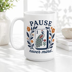 Op de afbeelding: Een witte keramische mok met een donkerblauwe grafische opdruk met de tekst PAUSE bovenaan en never mind... onderaan. In het midden staat een rechthoekige tegelillustratie van een pauw met het cijfer 1, geflankeerd door bloemrijke ranken.