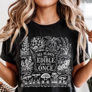 May include: A black t-shirt featuring a white botanical illustration of various poisonous plants and mushrooms. The central text reads, "ALL PLANTS ARE EDIBLE SOME ONLY ONCE," surrounded by labeled drawings of foxglove, oleander, and other toxic species.