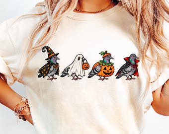 May include: A cream-colored t-shirt featuring a graphic print of four pigeons in Halloween costumes. The pigeons are dressed as a witch with a star-patterned hat and cape, a ghost, a pumpkin, and a vampire with a red-lined black cape.