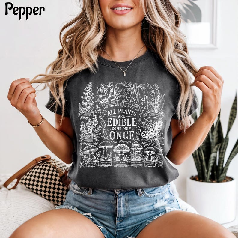 May include: A charcoal gray t-shirt featuring a white botanical graphic print of various mushrooms and plants. The text in the center reads, "ALL PLANTS ARE EDIBLE SOME ONLY ONCE," surrounded by detailed illustrations of toxic flora and fungi.