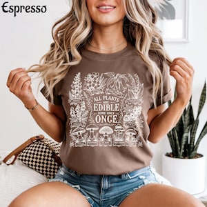 May include: A brown t-shirt featuring a white botanical graphic print of various plants and mushrooms. The central text reads, "ALL PLANTS ARE EDIBLE SOME ONLY ONCE," surrounded by detailed illustrations of flora with small labels underneath each item.