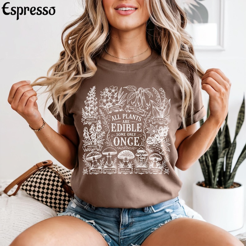 May include: A brown t-shirt featuring a white botanical graphic print of various plants and mushrooms. The central text reads, "ALL PLANTS ARE EDIBLE SOME ONLY ONCE," surrounded by detailed illustrations of flora with small labels underneath each item.