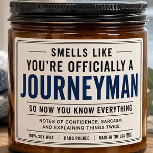May include: A brown glass jar candle with a black lid and a white label featuring the text: SMELLS LIKE YOU'RE OFFICIALLY A JOURNEYMAN SO NOW YOU KNOW EVERYTHING. NOTES OF CONFIDENCE, SARCASM, AND EXPLAINING THINGS TWICE. 100% SOY WAX, HAND POURED, MADE IN THE USA.