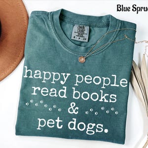 Op de afbeelding: Een blauw-sparrenkleurig t-shirt met korte mouwen en een ronde hals. Het shirt is voorzien van de witte tekst "happy people read books & pet dogs." met kleine pootafdrukken tussen de woorden. Een label is zichtbaar bij de halslijn.