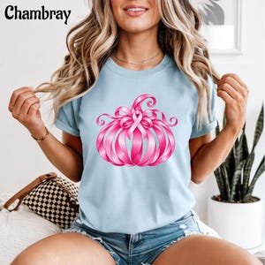 Op de afbeelding: Een lichtblauw t-shirt met een afbeelding van een pompoen gemaakt van roze linten, met een opvallend roze lint voor borstkankerbewustzijn in het midden. Het woord Chambray staat in zwarte letters linksboven in de afbeelding.