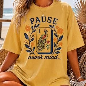 Op de afbeelding: Een mosterdgeel T-shirt met korte mouwen en een grafische print van een mahjongtegel met een pauwillustratie en het cijfer 1. Het ontwerp bevat de tekst PAUSE boven de tegel en never mind... eronder, omlijst door oranje bloemen en blauwe bladeren.
