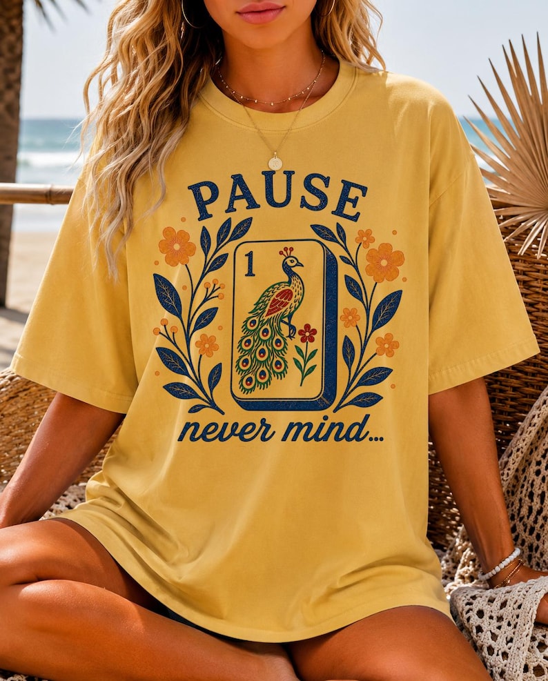 Op de afbeelding: Een mosterdgeel T-shirt met korte mouwen en een grafische print van een mahjongtegel met een pauwillustratie en het cijfer 1. Het ontwerp bevat de tekst PAUSE boven de tegel en never mind... eronder, omlijst door oranje bloemen en blauwe bladeren.