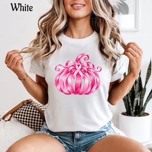 Op de afbeelding: Een wit t-shirt met korte mouwen en een afbeelding van een pompoen gemaakt van roze linten. Het ontwerp bevat een opvallend roze bewustzijnslint in het midden van de pompoen, waarbij krullende lintlussen de vorm van de pompoen vormen.