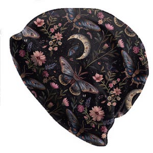 May include: A black beanie featuring a detailed pattern of moths, flowers, and crescent moons. The design incorporates shades of pink, blue, and cream against a dark background. The beanie is a soft, comfortable accessory.