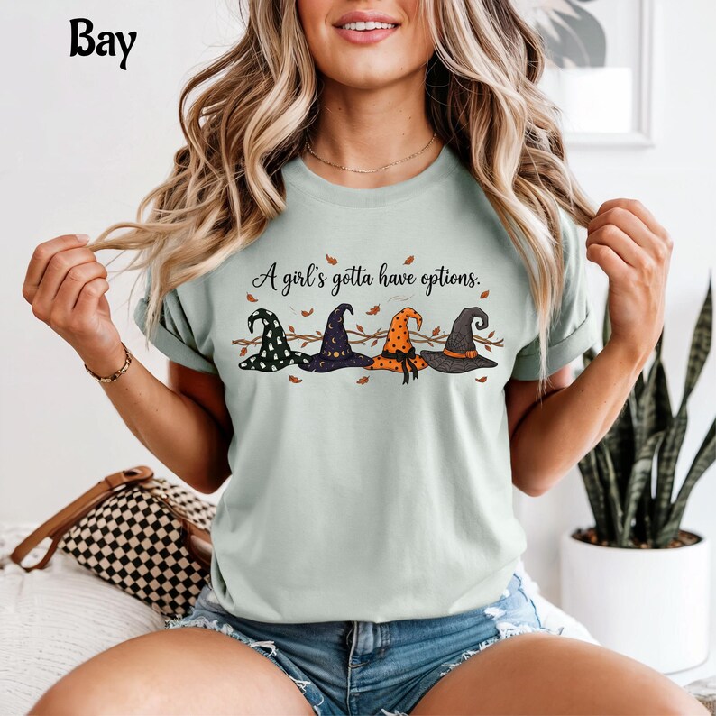 May include: A light green t-shirt featuring a graphic of four distinct witch hats in black, purple, and orange, with the text "A girl's gotta have options." printed above the hats, surrounded by small scattered autumn leaves and thin branches.