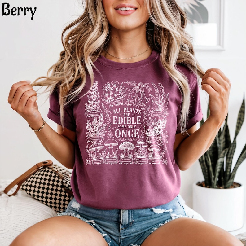 May include: A berry-colored short-sleeve t-shirt featuring a white graphic print of various botanical illustrations and mushrooms. The central text reads, "ALL PLANTS ARE EDIBLE SOME ONLY ONCE," surrounded by detailed drawings of plants and fungi.