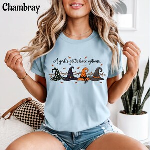 May include: A light blue crew neck t-shirt featuring a graphic of four distinct witch hats in black, purple, and orange. The text "A girl's gotta have options." is printed above the hats, accompanied by small brown leaf illustrations.