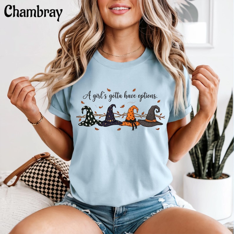 May include: A light blue crew neck t-shirt featuring a graphic of four distinct witch hats in black, purple, and orange. The text "A girl's gotta have options." is printed above the hats, accompanied by small brown leaf illustrations.